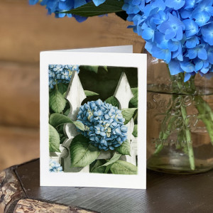white notecard of a painting of blue hydrangea and green leaves poking through a white fence on front