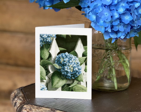 white notecard of a painting of blue hydrangea and green leaves poking through a white fence on front
