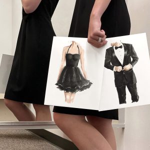 Lady holding two watercolor art prints of a lady in a little black dress and a man in a tuxedo